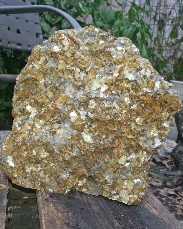 A large irregularly shaped golden rock sitting in front of a lawn chair, its jagged faces reflecting the sunlight in many different shades of yellow.