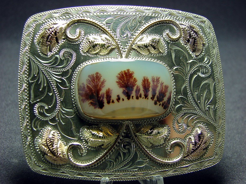 A small white agate cabochon with some brown dendrites that look like a row of trees. It has been set in an extremely ornate silver border.
