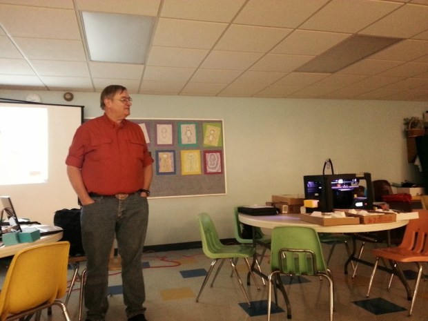 A man giving a presentation while standing next to a projector and a 3D printer.