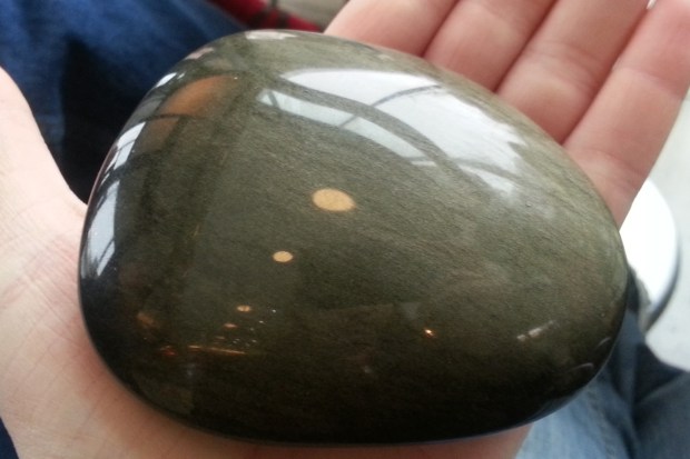 A dull gold oval-shaped stone resting comfortably in a person's palm.  It is highly polished and reflects the overhead lights, and appears to have depth.