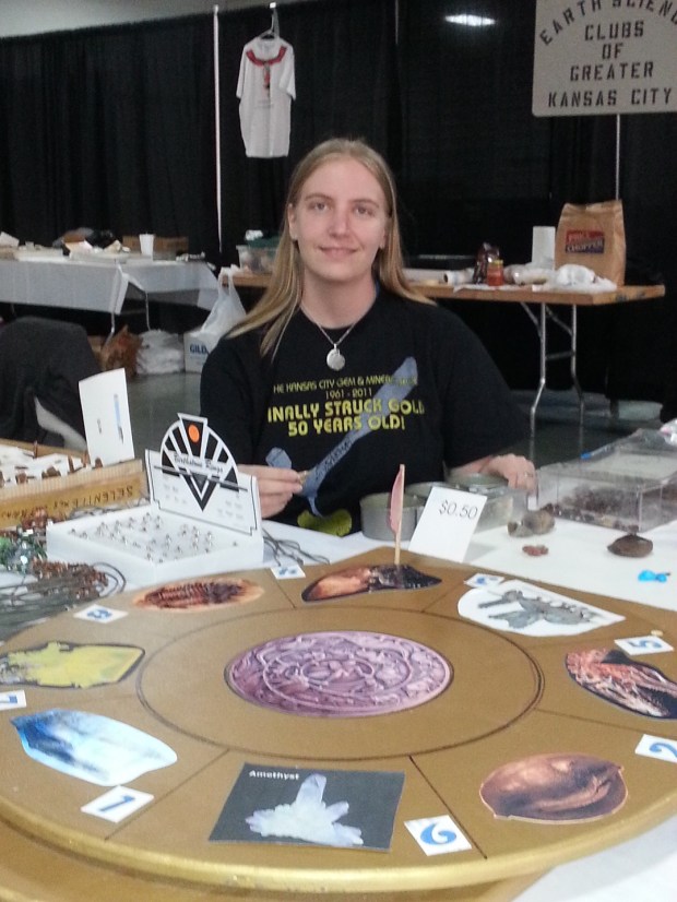 A smiling young woman sitting behind a large wooden spinner with eight sections. She is wearing an ammonite necklace and a shirt proclaiming that the Kansas City Gem and Mineral Show "finally struck gold" and is 50 years old.