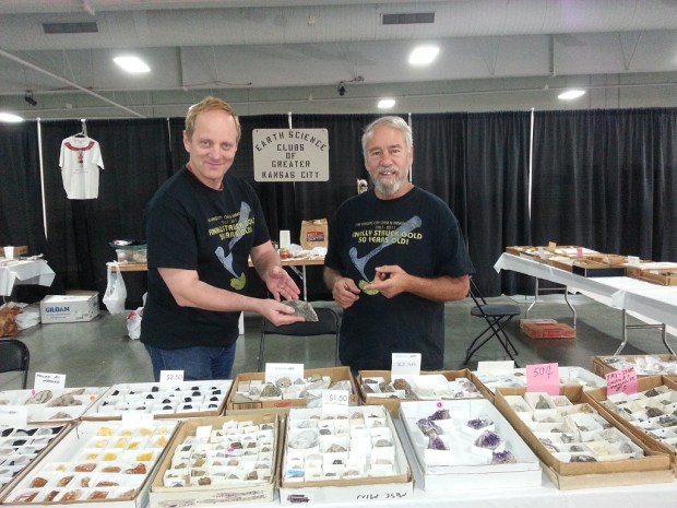 Two men holding rocks and standing in front of a table covered in boxes of colorful crystals. Behind them is a sign that reads, 