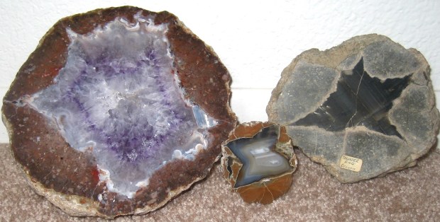 Three round geodes in small, medium, and large. The smallest one is a flat slab with a blue inside and brown outside. The medium one is a hemisphere that's black on the inside and gray on the outside. The largest one is a hemisphere that is purple amethyst crystals on the inside and brown on the outside.
