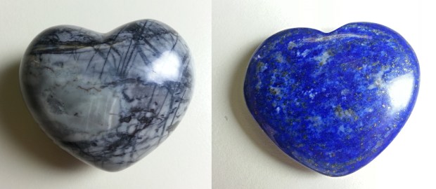 Two shiny stones carved into heart shapes. One is gray with black cross-hatched lines and the other is blue with small white swirls and gold sparkles. 