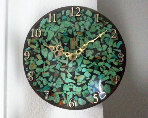 A round clock with green turquoise chips floating in resin on a black background. It has ornate gold hands and numbers that complement the gold flecks in the turquoise.
