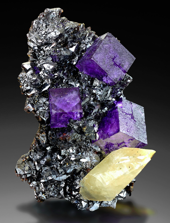 A silver sphalerite matrix with some purple fluorite crystals and a yellow calcite crystal on it.